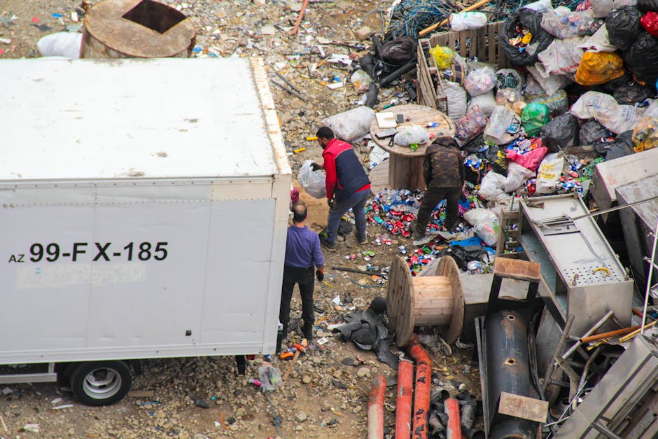 A street scene featuring a modern waste collection truck parked in front of multi-storey buildings with varied architectural styles, including brick facades, arched windows, and decorative rooflines. 